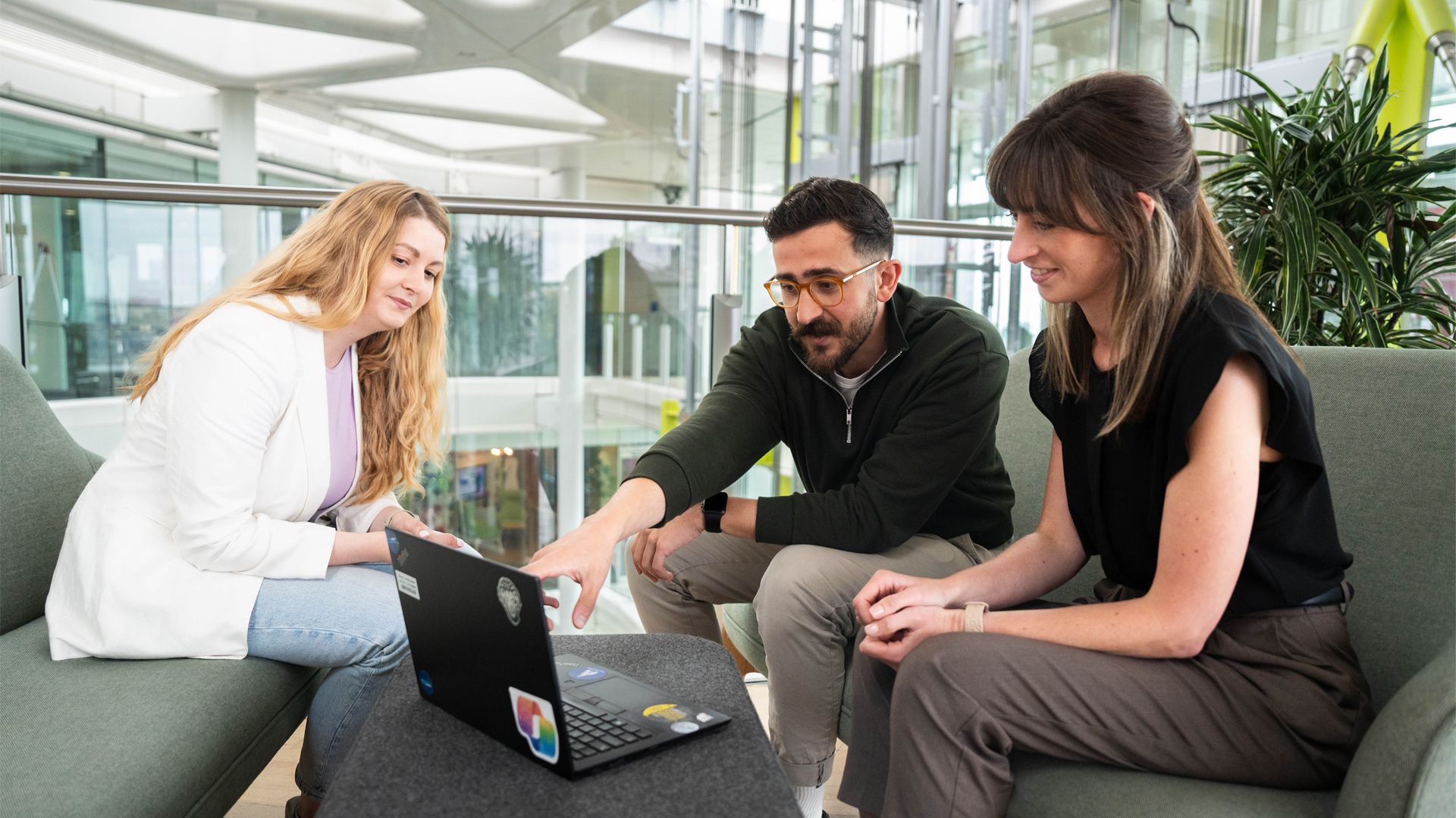 Three people seated on two couches in a modern office, collaborating around a laptop on a table. One person points at the screen while the others observe, with large windows and indoor plants in the background.