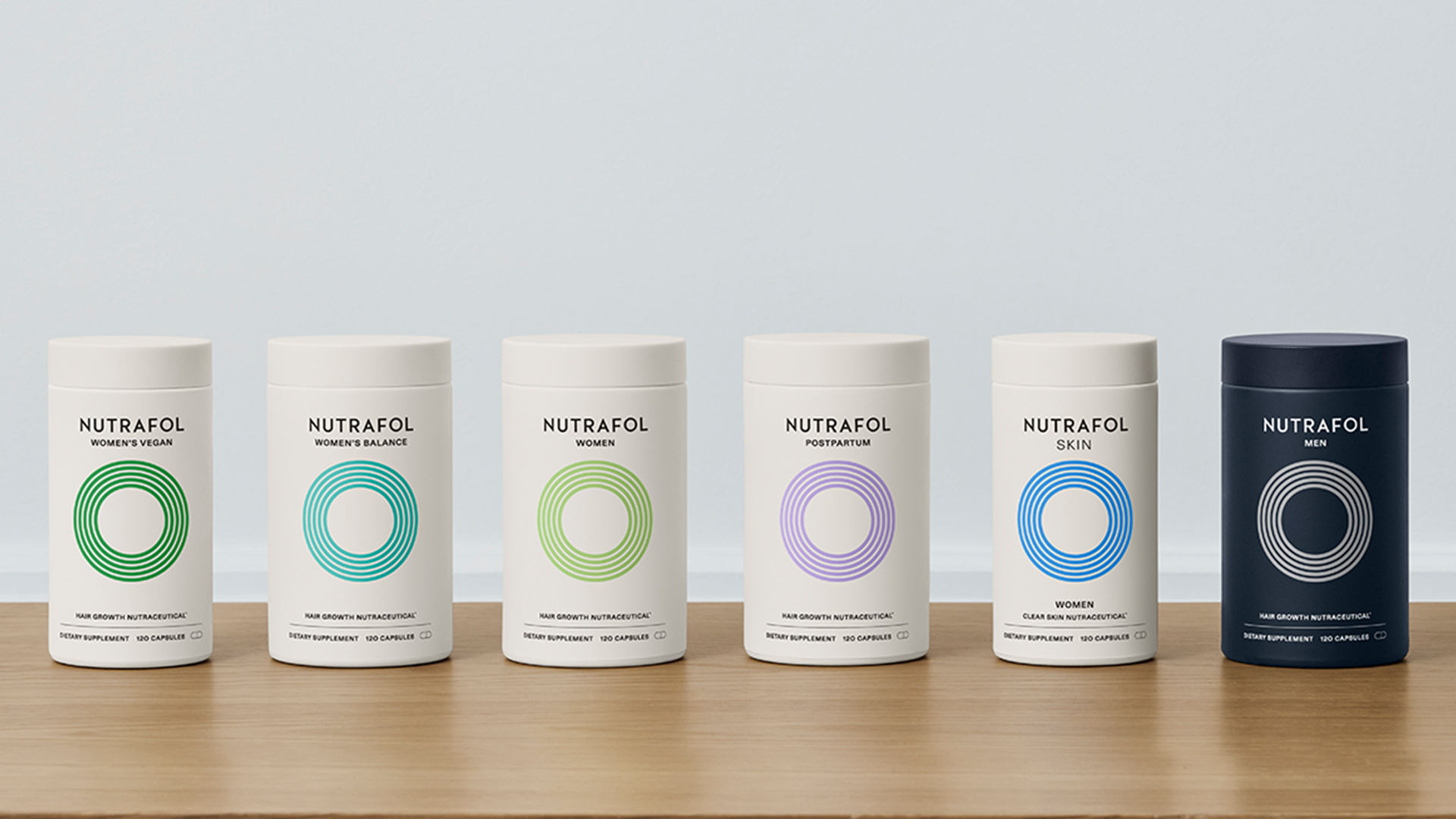 Six Nutrafol products positioned in a row on a wooden table – five are in white packaging and one appears in black.