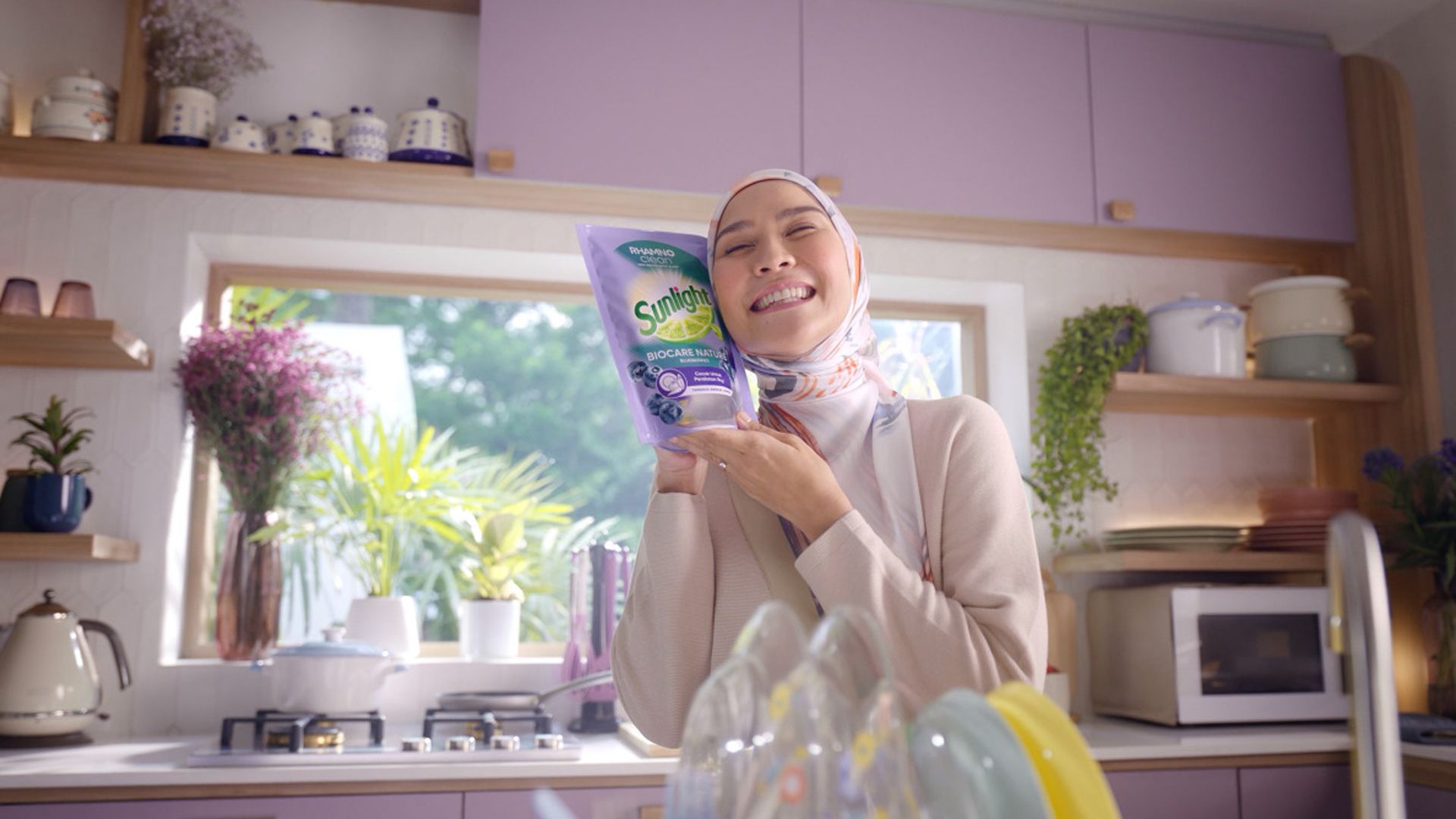 Someone standing in a kitchen, holding a pouch of Sunlight up against their face, smiling widely. In the background are shelves with storage jars and cooking pots on them, a microwave and kettle. Behind is a window with plant pots and a vase of flowers. In the foreground are some plates draining near a sink.