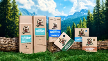 Dr. Squatch grooming products – including shampoo, conditioner, deodorant, and soaps – arranged on logs and grass in a natural outdoor setting with trees, mountains, and a blue sky in the background.