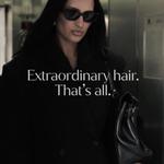 A person wearing a dark coat and sunglasses stands indoors holding a black handbag, with long dark hair visible. Overlaid text reads: “Extraordinary hair. That’s all.”