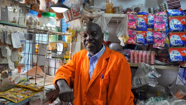 A smiling shopkeeper wearing an orange coat stands inside a small convenience store. Behind him, shelves are stocked with various products, including ‘OMO’ and ‘Sunlight’.
