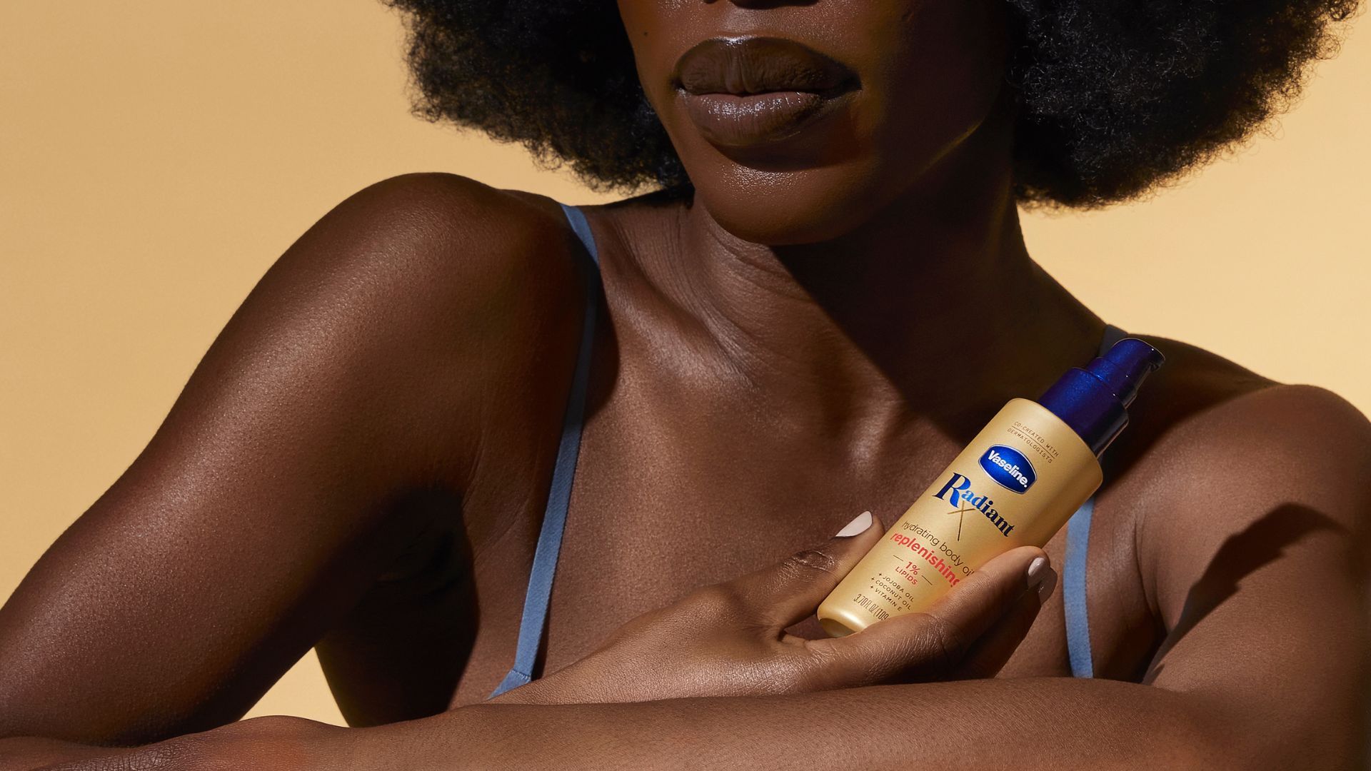 A Black woman holds Vaseline Radiant X Replenishing Hydrating Body Oil. Her arms, chest and lower face are visible.