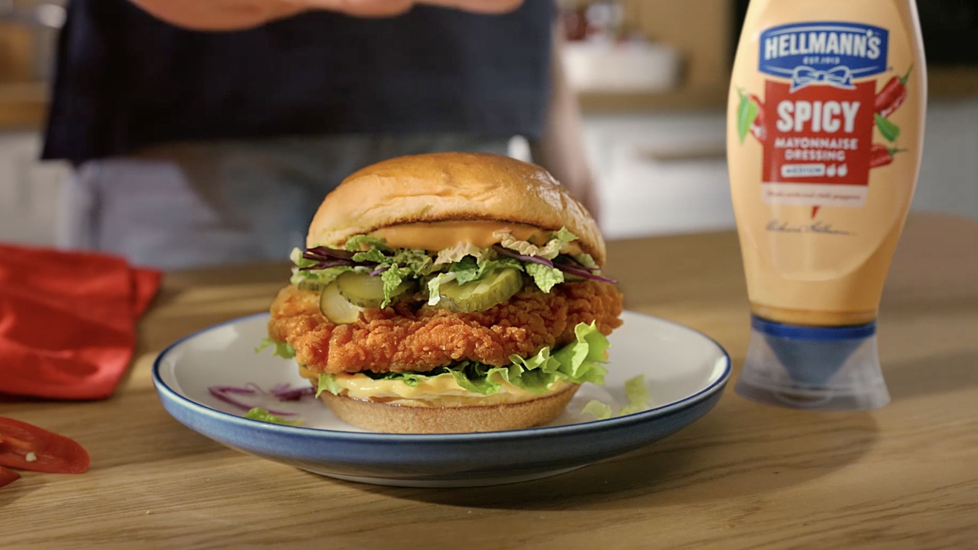 Close-up of a crispy fried chicken sandwich on a plate, layered with lettuce, pickles, and avocado slices inside a soft bun. A bottle of Hellmann’s Spicy Mayonnaise Dressing is placed to the right, and a red napkin is partially visible on the left. The setting appears to be a kitchen countertop with warm lighting.