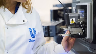 A close-up photo of a scientist in a Unilever-branded white lab coat, looking at a large digital microscope.