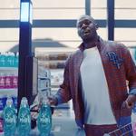 Usain Bolt standing at a checkout counter in a brightly lit store, wearing a patterned jacket and white shirt, holding a bottle of Persil detergent. Shelves in the background are stocked with colourful bottles of cleaning products in pink, blue, and turquoise.