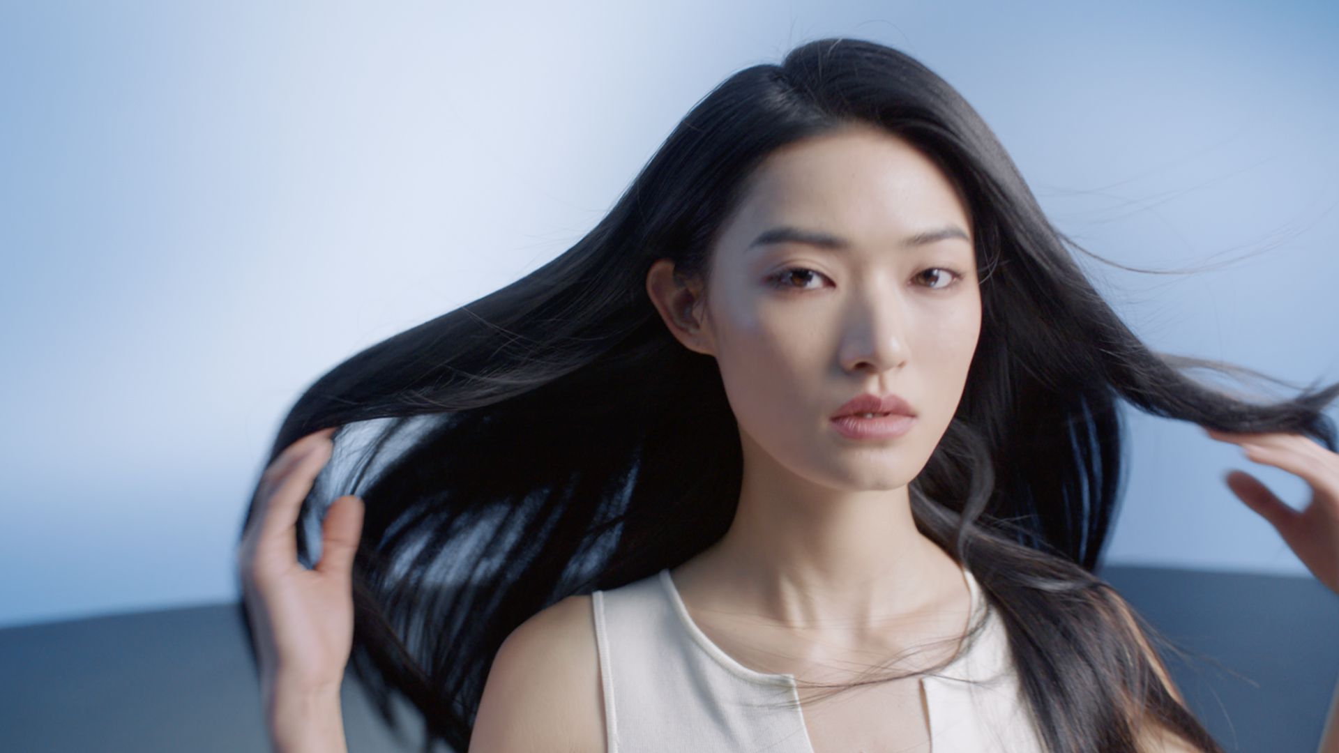 Person wearing a sleeveless top with long, shiny hair flowing outward against a soft blue background.