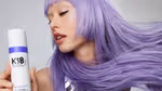 A woman with long violet hair and winged eyeliner holding a bottle of K18 Airwash Dry Shampoo.