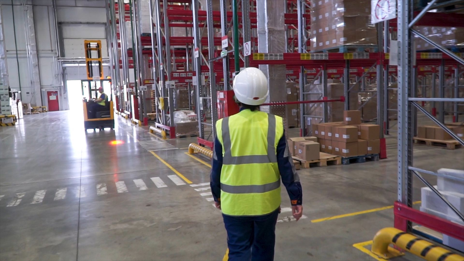Female warehouse worker in high-visibility vest and hard hat walking towards some boxes on shelves
