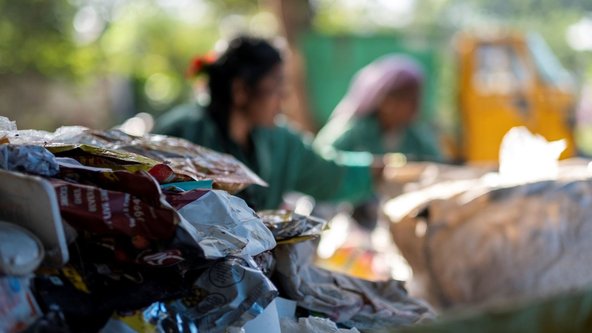 A close-up of a pile of discarded plastic packaging, with two workers sorting waste in the blurred background.