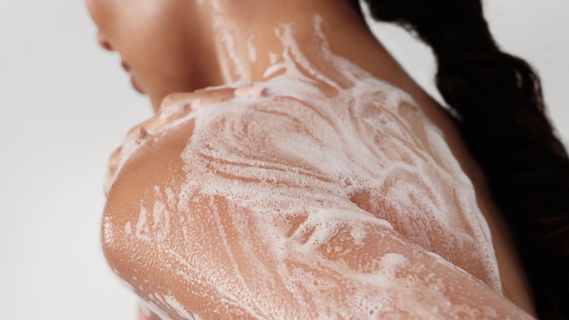 Back view of a woman with her hair in a ponytail whose naked back and shoulders are lathered with Dove’s foaming body wash   
