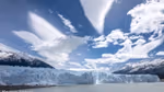 View of the terminus of a glacier as seen from sea level with mountains in the background and a bright, sunny sky
