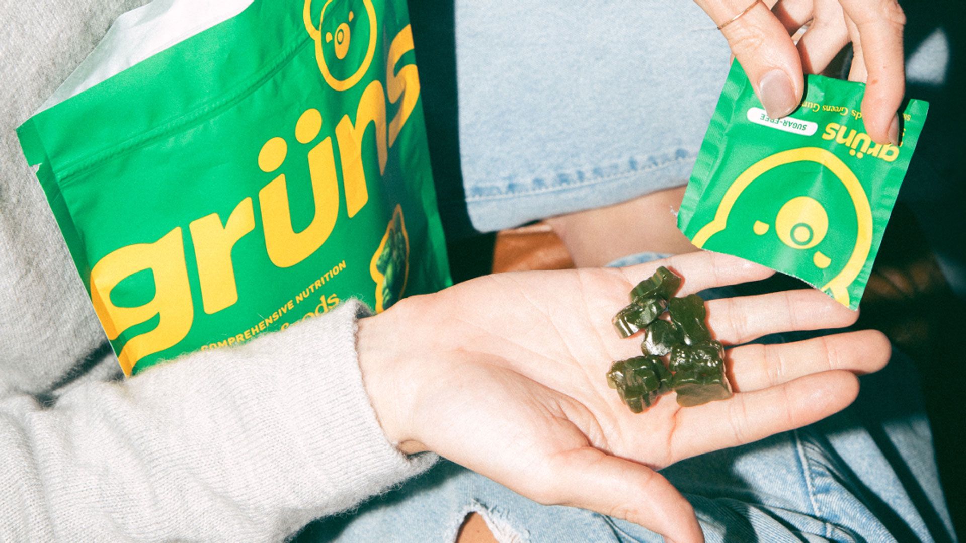Green gummy supplements resting in an open hand, with more gummies being poured from a small Grüns packet; a larger Grüns pouch is visible in the background.