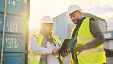 Two workers using a digital tablet discussing ecommerce, logistics, supply chain and shipping. Freight and cargo in the background.