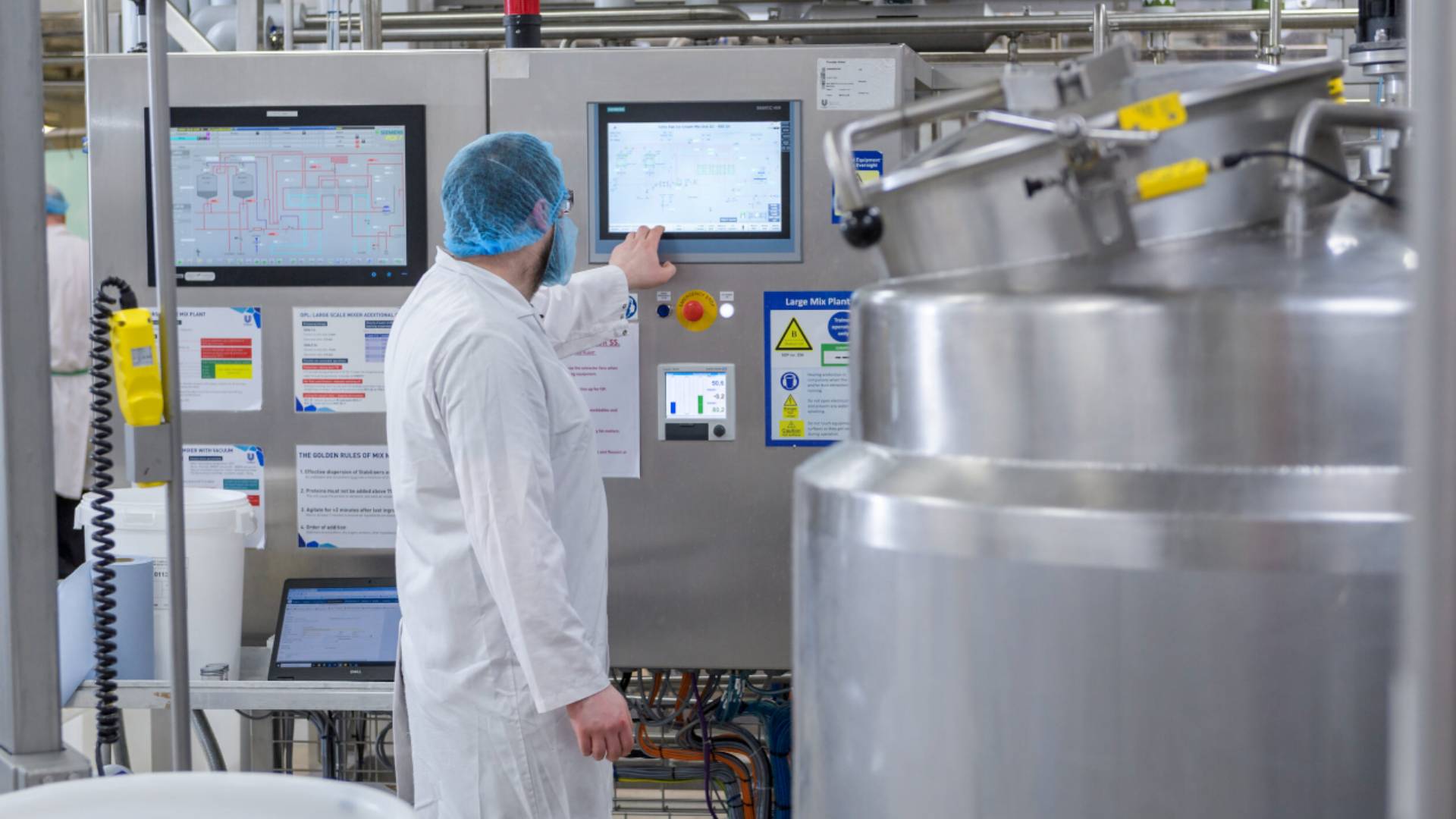 Ice cream factory worker in a white lab coat and hairnet attending a machine that’s part of the manufacturing process 