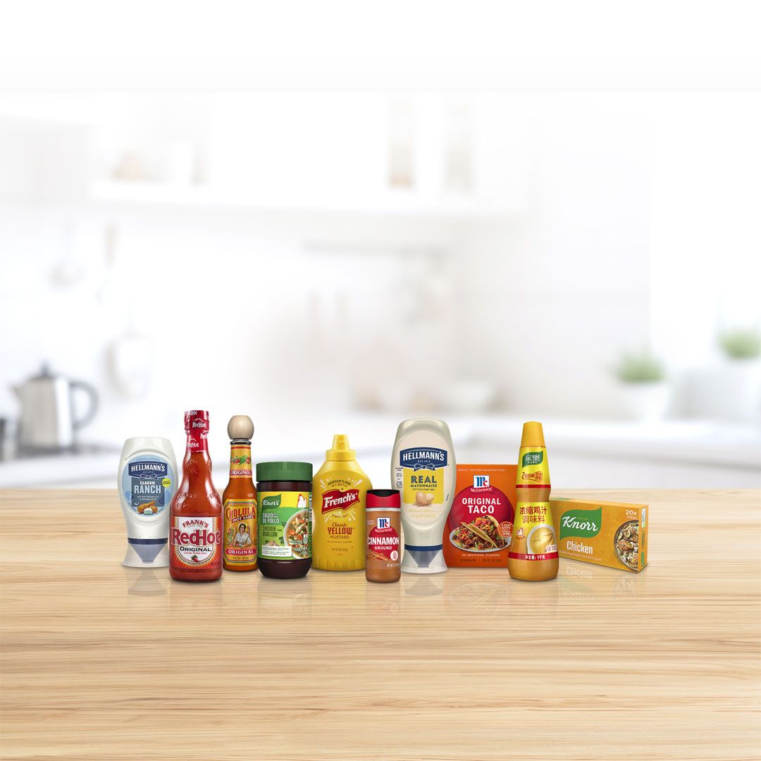 A selection of well known food and condiment brands, including Hellmann’s mayonnaise, Frank’s RedHot, Cholula Hot Sauce, Knorr seasonings, French’s mustard, and condiments, arranged side by side on a wooden table.