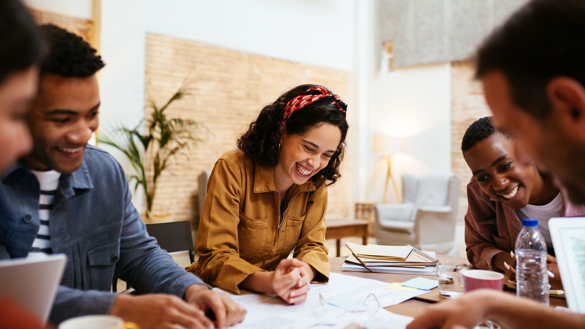 A group of people sitting around a table in a bright, informal workspace, looking over documents and laptops during a collaborative meeting.