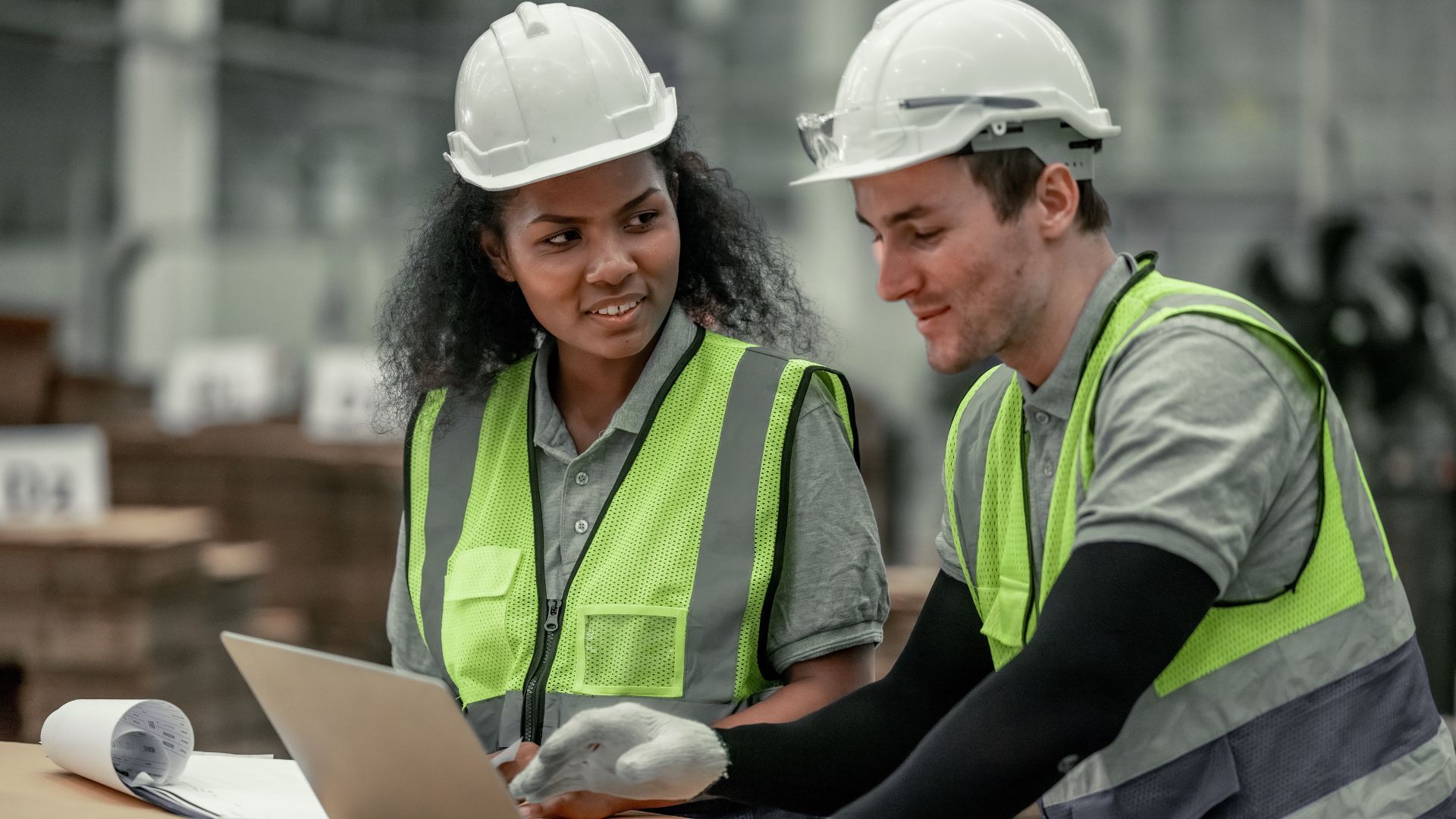 Two people in hard hats in a factory chatting and looking at a laptop