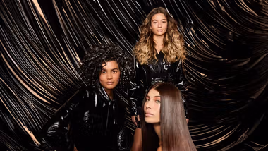 Three women with shiny hair, one blonde waves, one dark curls, one dark glossy. Photo is an ad for TRESemmé Lamellar Shine.