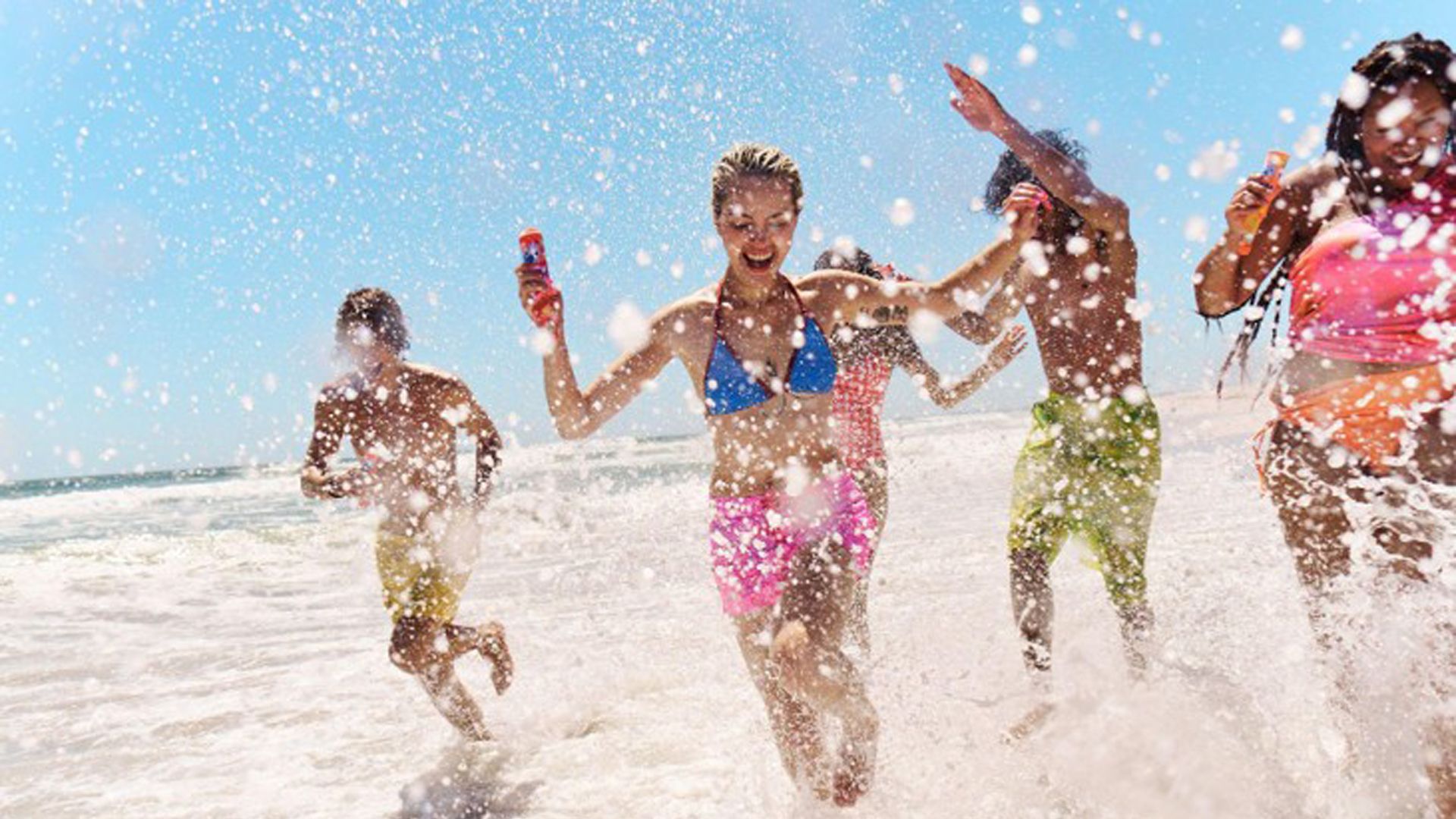 Five people running through shallow water at a beach, splashing waves around them under a clear blue sky. They are dressed in colourful swimwear and appear to be enjoying a sunny day.