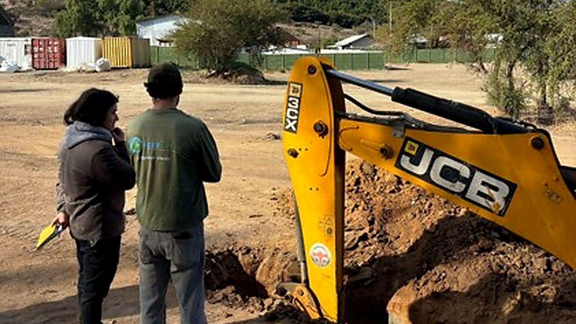 Two workers inspect an excavator digging a trench on a construction site.