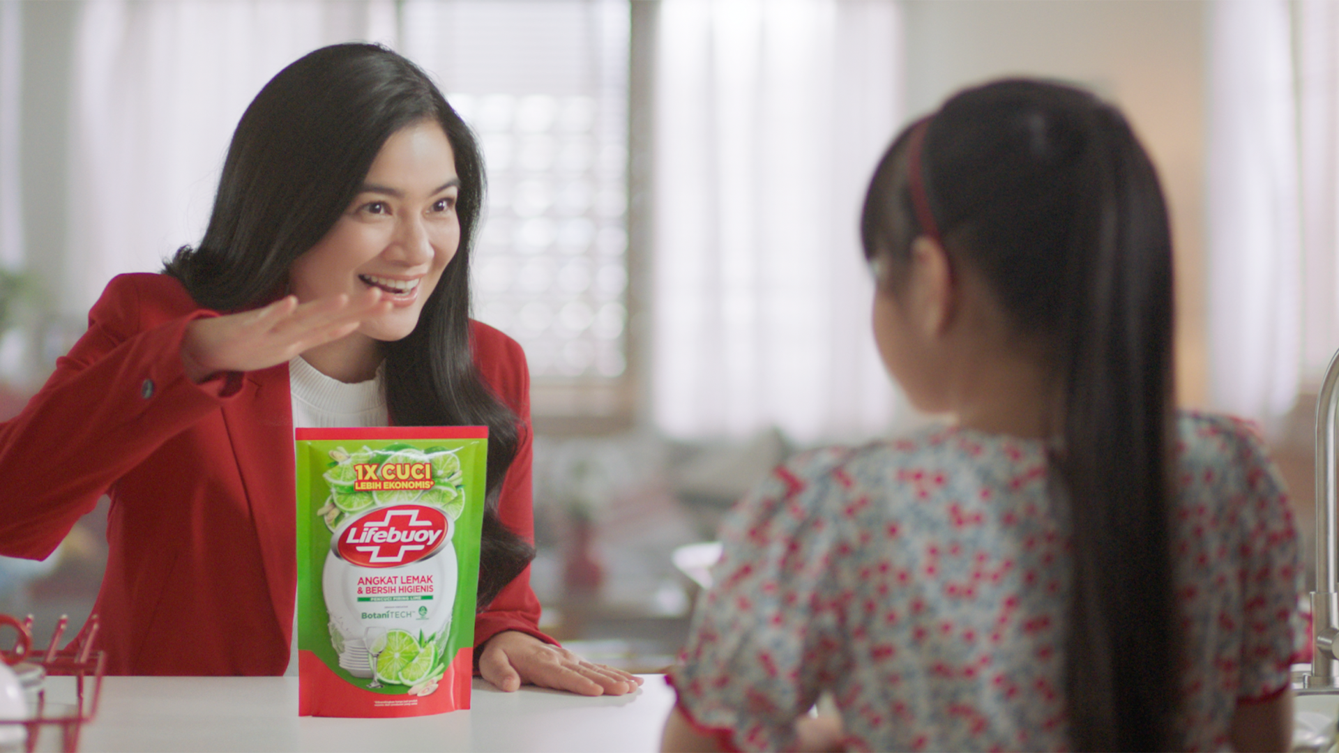 A woman in a red jacket, stands with a pack of Lifebuoy dishwash liquid. A girl is in the foreground facing her.