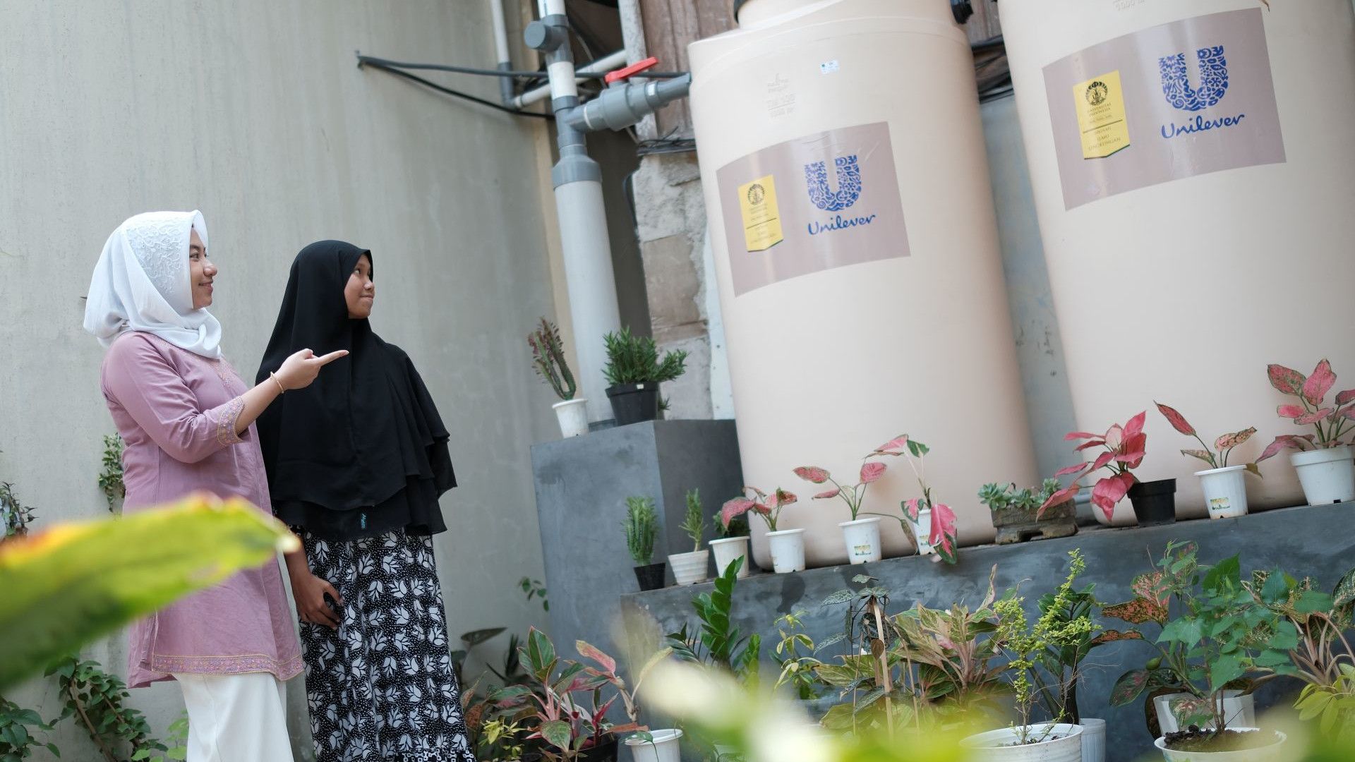 A photograph of two female students observing a Unilever rain harvesting installation at an Islamic boarding school in Cikarang, West Java, Indonesia, with green vegetation in the foreground.