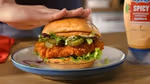A chicken burger on a plate on a wooden table. To the right of the plate is a bottle of Hellmann’s spicy mayo. A hand is pressing down lightly on top of the burger.