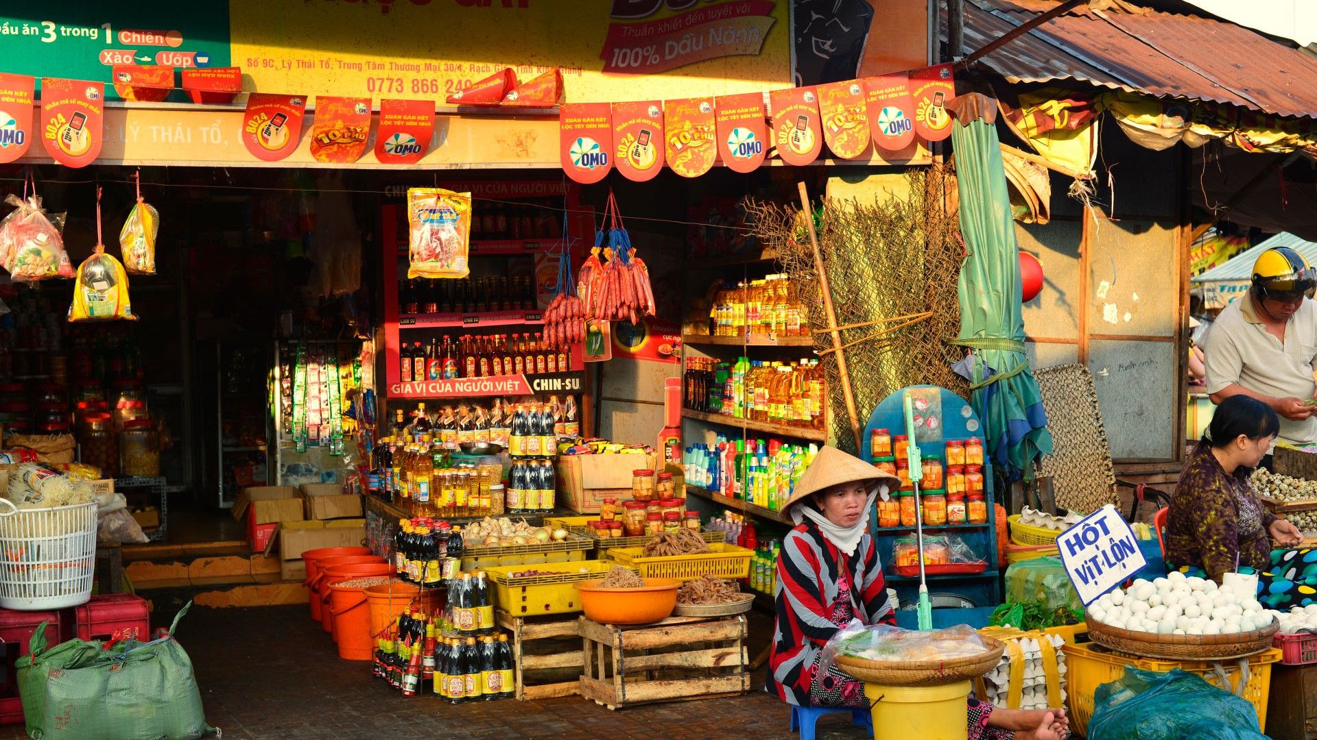 A vibrant outdoor market stall showcasing a variety of goods for sale. The stall is stocked with colourful products, neatly displayed on shelves and in baskets. In the foreground is a woman wearing a traditional conical hat and a striped shirt sitting next to a basket of goods.