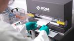 A laboratory technician wearing green gloves places a sample sheet into an RDM Test Equipment machine. The background shows additional laboratory apparatus and tools.