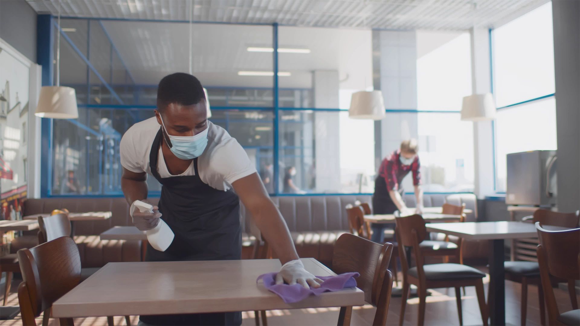 Two people wearing facemasks wiping down tables in a restaurant