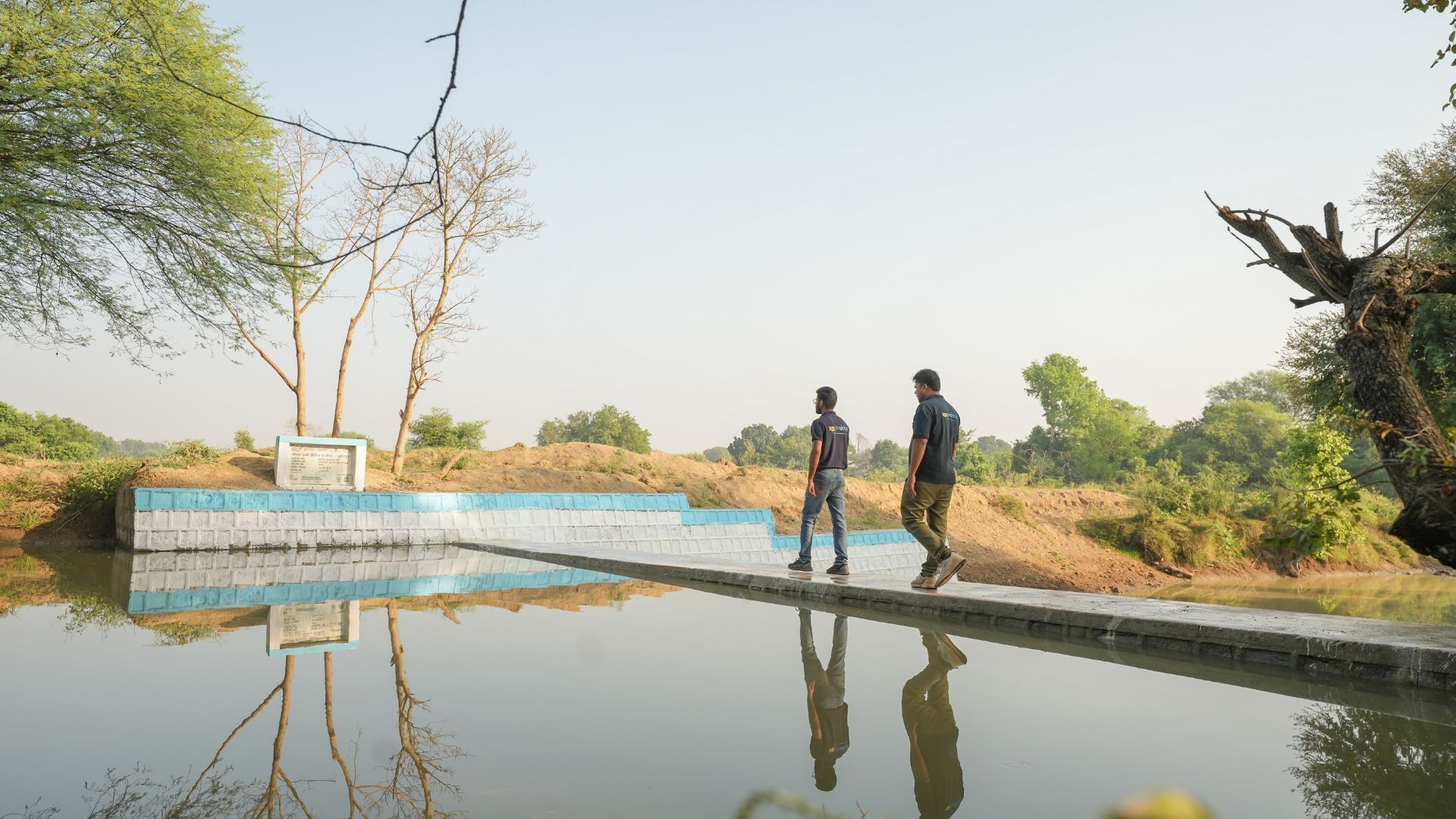 Two workers inspect a concrete water structure beside a calm, reflective reservoir in a rural landscape