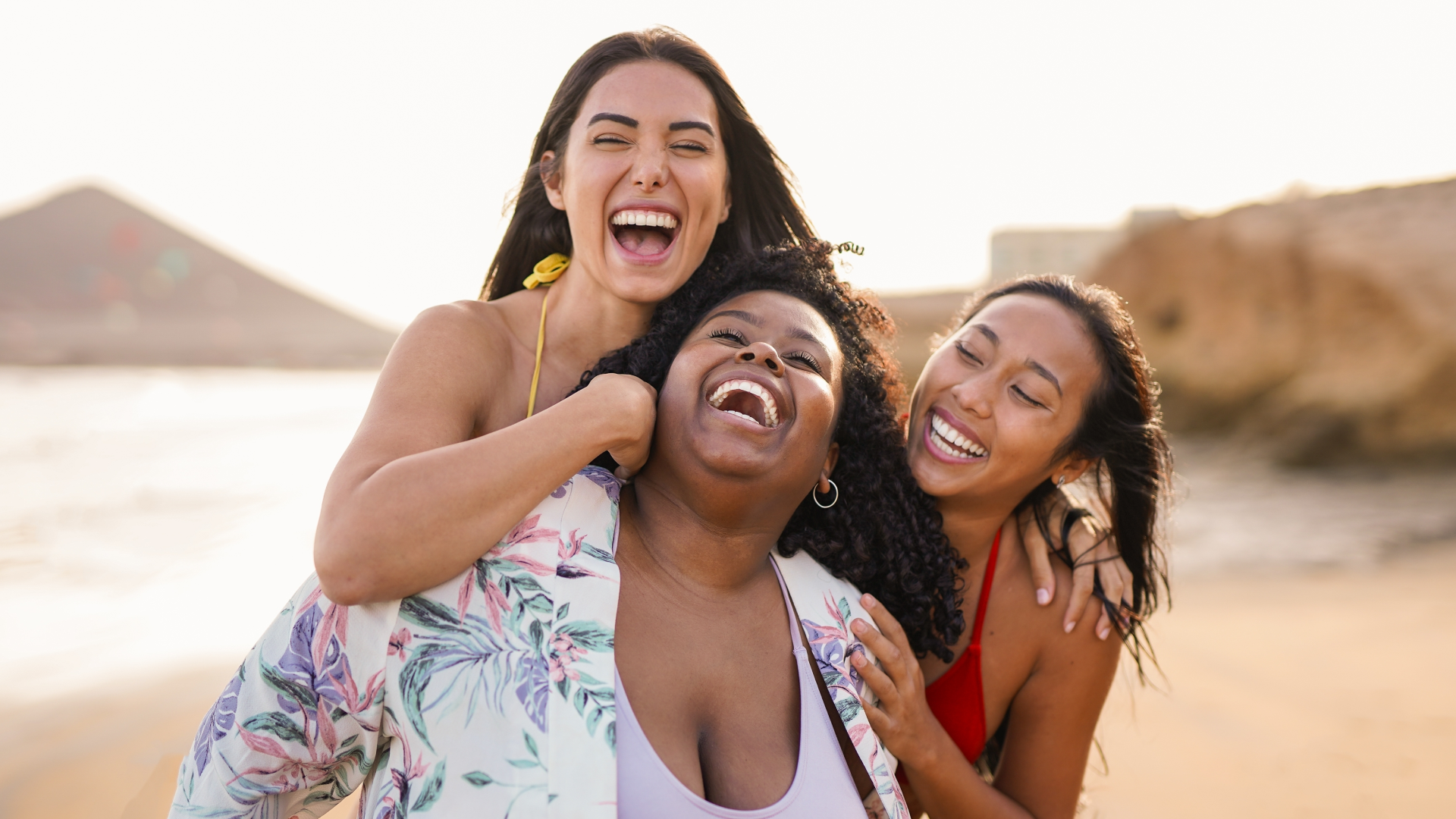 Three people standing close together on a sandy beach, wearing colorful swimwear and a floral cover-up, with rocky cliffs and ocean in the background.