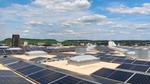 Solar panels cover a large rooftop overlooking a city and green hills.