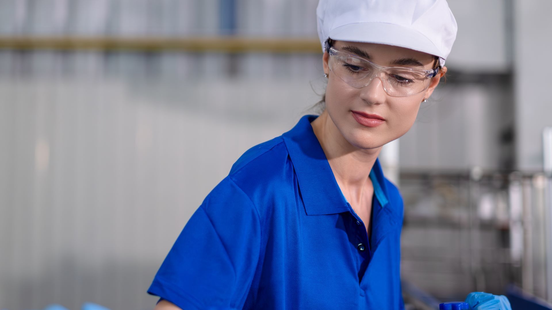 A worker wearing a blue uniform, a white cap, and clear safety glasses in an industrial setting.