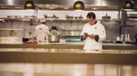 A chef consulting his phone in a professional kitchen while another chef works in the background