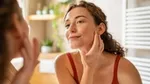 Woman in a bathroom, smiling at her reflection in the mirror. She has her hand to her face and is wearing a red top.