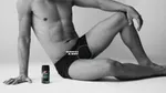 A bare-chested man in black pants sits on the floor in a relaxed pose, one knee raised with his wrist resting on it. A can of Lynx body spray is next to him. A “SCRATCH & SNIFF” label points to his shorts. The image is in black and white.