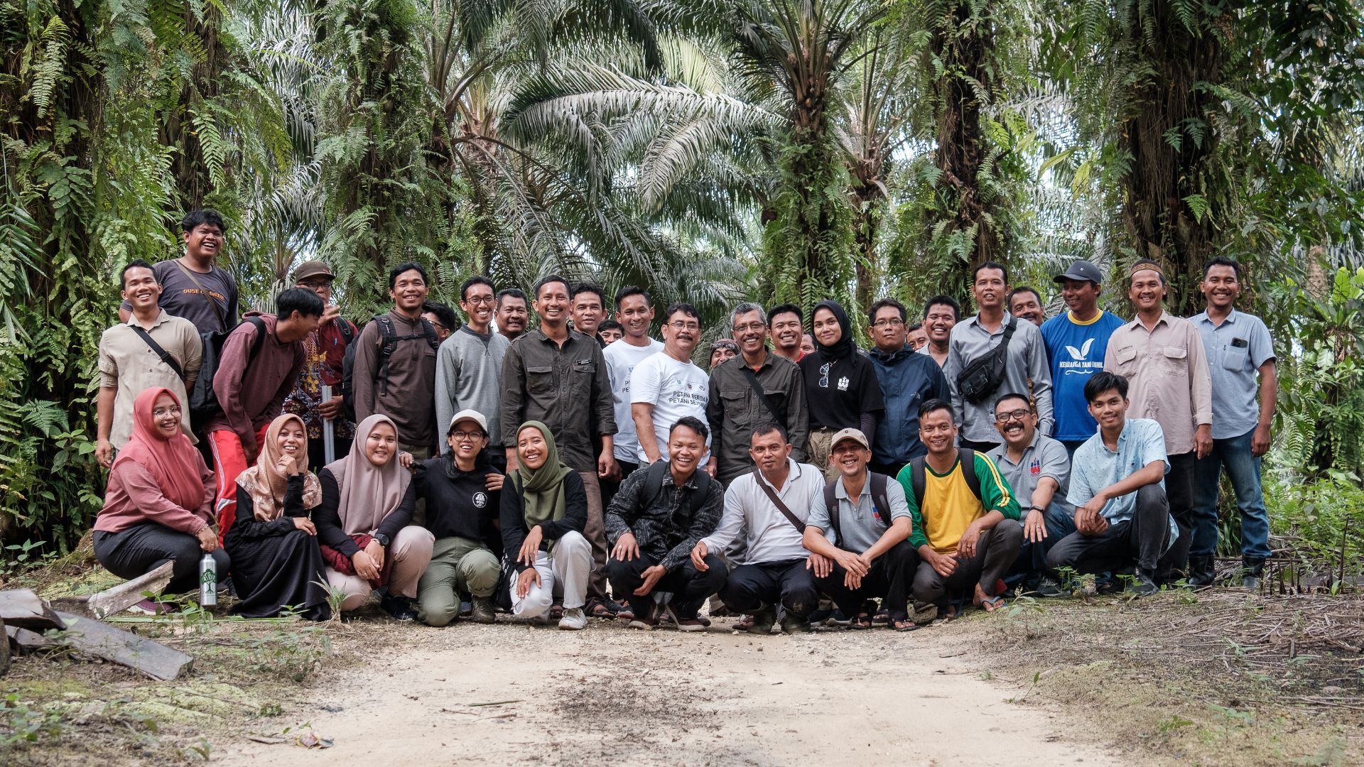 A photograph of a large group of male and female smallholder farmers, some standing, some crouched down, with palm trees in the background, in Riau, Indonesia.