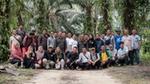 A photograph of a large group of male and female smallholder farmers, some standing, some crouched down, with palm trees in the background, in Riau, Indonesia.