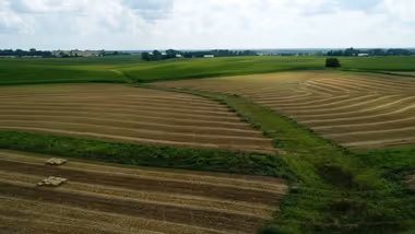Countryside aerial view showing neatly mowed fields with hay bales and large areas of green grass, under a partly cloudy sky.