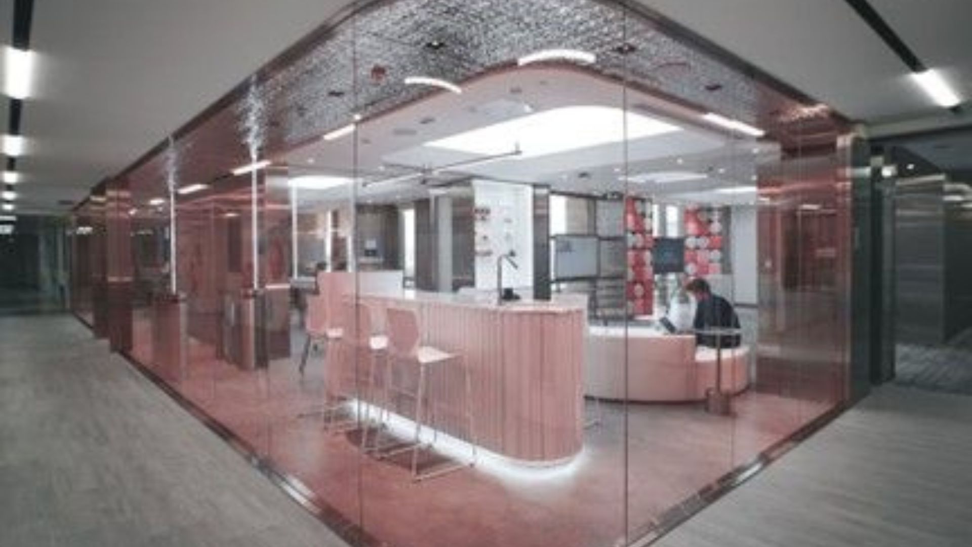 The interior of an office building, viewed from a corridor looking into a meeting room with various seating options and large TV screens. A person is sitting and working on a laptop
