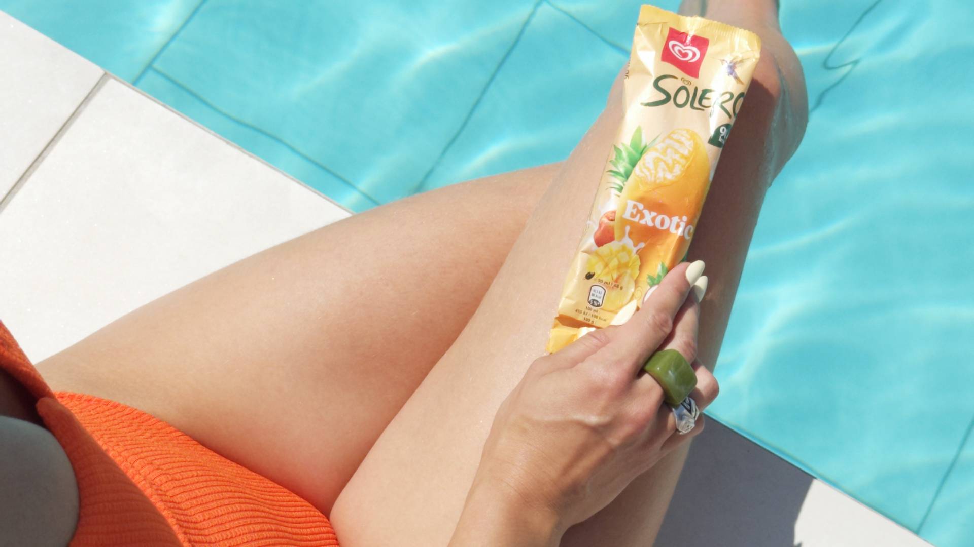 A woman in an orange swimsuit is sitting by the edge of a swimming pool holding a wrapped Solero ice cream